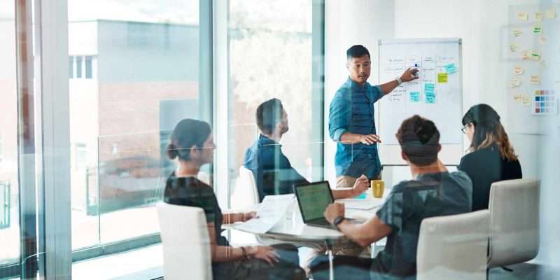 Boost your business Team meeting in a glass walled office. Person using post-its on a whiteboard to explain their idea