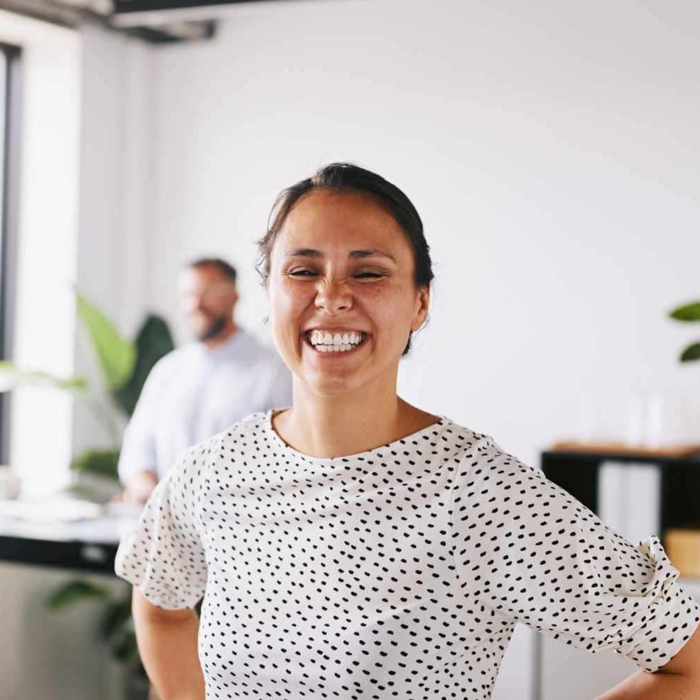 Woman in an office smiling broadly at camera in a casual pose wearing a white t-shirt with black spots